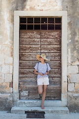 Image showing Beautiful young female tourist woman standing in front of vinatage wooden door and textured stone wall at old Mediterranean town, smiling, holding, smart phone to network on vacationes