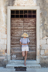 Image showing Beautiful young female tourist woman standing in front of vinatage wooden window and textured stone wall at old Mediterranean town, smiling, holding, using smart phone to network on vacationes