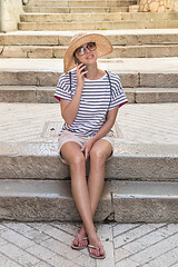 Image showing Beautiful young tourist woman at summer vacations, sitting on old stone steps of old medieval Mediterranean costal town, smiling, talking on phone with her loved ones at home