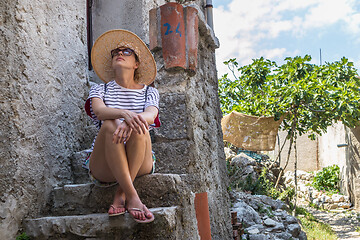 Image showing Beautiful female tourist wearing big straw sun hat and shorts sitting and relaxing on old stone house stairs during summer travel on Mediterranean cost on hot summer day