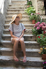 Image showing Beautiful female tourist wearing big straw sun hat and shorts sitting and relaxing on old stone house stairs during summer travel on Mediterranean cost on hot summer day