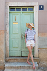 Image showing Beautiful young female tourist woman wearing sun hat, standing and relaxing in shade in front of vinatage wooden door in old Mediterranean town while sightseeing on hot summer day