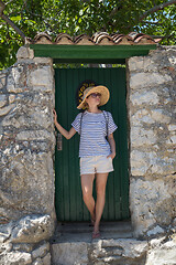 Image showing Beautiful young female tourist woman wearing sun hat, standing and relaxing in shade in front of vinatage wooden door in old Mediterranean town while sightseeing on hot summer day