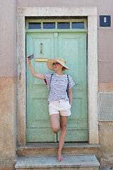 Image showing Beautiful young female tourist woman wearing big straw hat, taking self portrait selfie, standing in front of turquoise vinatage wooden door and textured stone wall at old Mediterranean town