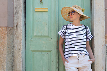 Image showing Beautiful young female tourist woman wearing sun hat, standing and relaxing in shade in front of vinatage wooden door in old Mediterranean town while sightseeing on hot summer day