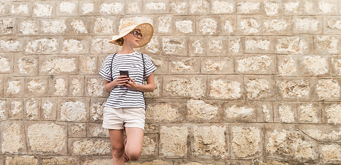 Image showing Beautiful young female tourist woman standing in front of old textured stone wall at old Mediterranean town, smiling, holding, smart phone to network on vacationes. Copy space