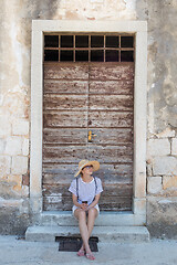 Image showing Beautiful young female tourist woman sitting and resting on vinatage wooden doorstep and textured stone wall at old Mediterranean town, smiling, holding, using smart phone to network on vacationes