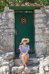 Image showing Beautiful young female tourist woman sitting and resting on vinatage wooden doorstep and textured stone wall at old Mediterranean town, smiling, holding, using smart phone to network on vacationes