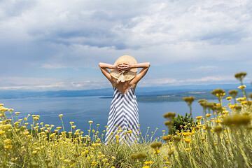 Image showing Rear view of young woman wearing striped summer dress and straw hat standing in super bloom of wildflowers, relaxing while enjoing beautiful view of Adriatic sea nature, Croatia