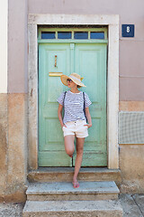 Image showing Beautiful young female tourist woman wearing sun hat, standing and relaxing in shade in front of vinatage wooden door in old Mediterranean town while sightseeing on hot summer day