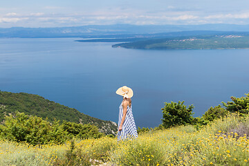 Image showing Rear view of young woman wearing striped summer dress and straw hat standing in super bloom of wildflowers, relaxing while enjoing beautiful view of Adriatic sea nature, Croatia