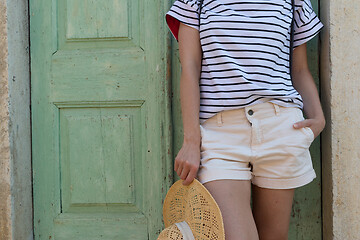 Image showing Fashinable torso portrait of woman holding sun hat, standing and relaxing in shade in front of turquoise vinatage wooden door in old Mediterranean town while sightseeing on hot summer day