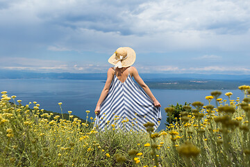 Image showing Rear view of young woman wearing striped summer dress and straw hat standing in super bloom of wildflowers, relaxing while enjoing beautiful view of Adriatic sea nature, Croatia