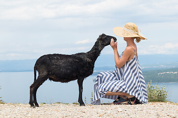 Image showing Young attractive female traveler wearing striped summer dress and straw hat squatting, feeding and petting black sheep while traveling Adriatic coast of Croatia