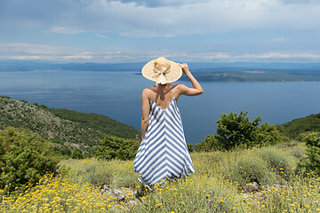 Image showing Rear view of young woman wearing striped summer dress and straw hat standing in super bloom of wildflowers, relaxing with hands up to the sky, enjoing beautiful view of Adriatic sea nature, Croatia