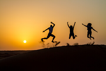 Image showing Silhouette of happy traveling people jumping on sand dune and throwing sand in the air in golden sunset hour