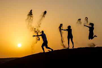 Image showing Silhouette of happy traveling people jumping on sand dune and throwing sand in the air in golden sunset hour