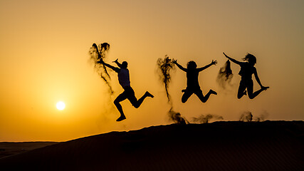 Image showing Silhouette of happy traveling people jumping on sand dune and throwing sand in the air in golden sunset hour