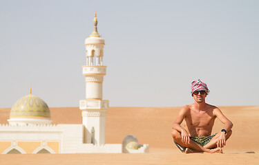 Image showing Man traveling in Oman, topless wearing a turban and shorts, sitting on send dune in fron of mosque in desert Oman