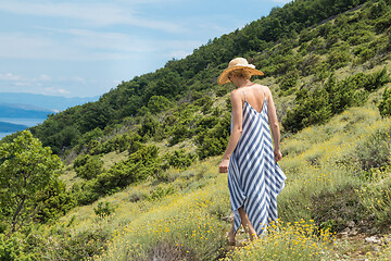 Image showing Young woman wearing striped summer dress and straw hat walking among super bloom of wildflowers, relaxing while enjoing beautiful nature of of Adriatic sea coastal nature of Croatia