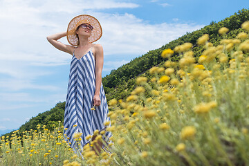 Image showing Young woman wearing striped summer dress and straw hat standing in super bloom of wildflowers, relaxing while enjoing beautiful nature of of Adriatic sea coastal nature of Croatia.