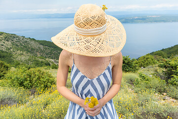 Image showing Young woman wearing striped summer dress and straw hat standing in super bloom of wildflowers, holding bouquet of yellow flowers in beautiful nature of Adriatic sea coastal nature of Croatia