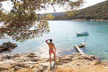 Image showing Rear view of man wearing red speedos tanning and realaxing on wild cove of Adriatic sea on a beach in shade of pine tree.