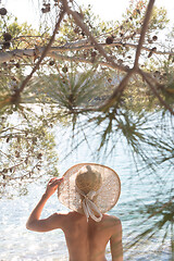 Image showing Rear view of topless beautiful woman wearing nothing but straw sun hat realaxing on wild coast of Adriatic sea on a beach in shade of pine tree.