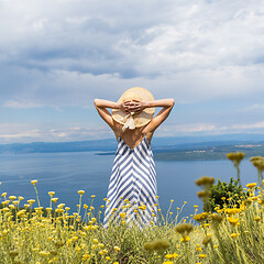 Image showing Rear view of young woman wearing striped summer dress and straw hat standing in super bloom of wildflowers, relaxing while enjoing beautiful view of Adriatic sea nature, Croatia