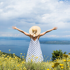 Image showing Rear view of young woman wearing striped summer dress and straw hat standing in super bloom of wildflowers, relaxing with hands up to the sky, enjoing beautiful view of Adriatic sea nature, Croatia