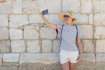 Image showing Beautiful young female tourist woman wearing big straw hat, taking self portrait selfie, standing in front of old textured stone wall at old Mediterranean town