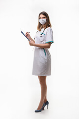 Image showing Slender nurse in medical dress and medical mask records data on white background