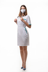 Image showing A girl in medical clothes and a mask holds a stethoscope on a white background