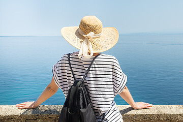 Image showing Rear view of woman traveler wearing straw summer hat and backpack,leaning against a stone wall looking at big blue sea and islands in on the horizon.