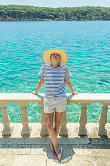 Image showing Rear view of woman wearing straw summer hat ,leaning against elegant old stone fence of coastal villa, relaxing while looking at blue Adriatic sea, on Losinj island Croatia.