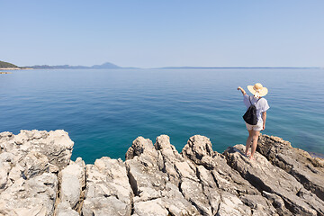 Image showing Woman traveler wearing straw summer hat and backpack, standing at edge of the rocky cliff looking and pointing at big blue sea and islands in on the horizon