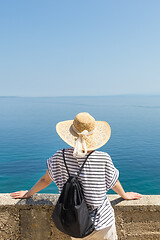 Image showing Rear view of woman traveler wearing straw summer hat and backpack,leaning against a stone wall looking at big blue sea and islands in on the horizon.