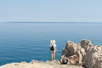 Image showing Woman traveler wearing straw summer hat and backpack, standing at edge of the rocky cliff looking at big blue sea and islands in on the horizon