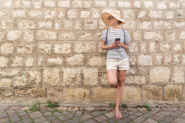 Image showing Beautiful young female tourist woman standing in front of old textured stone wall at old Mediterranean town, smiling, holding, smart phone to network on vacationes. Copy space