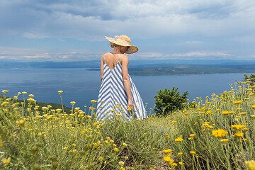 Image showing Rear view of young woman wearing striped summer dress and straw hat standing in super bloom of wildflowers, relaxing while enjoing beautiful view of Adriatic sea nature, Croatia