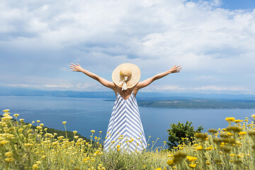 Image showing Rear view of young woman wearing striped summer dress and straw hat standing in super bloom of wildflowers, relaxing with hands up to the sky, enjoing beautiful view of Adriatic sea nature, Croatia