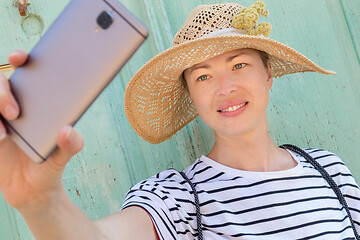 Image showing Beautiful young female tourist woman wearing big straw hat, taking self portrait selfie, standing in front of vinatage turquoise wooden door at old Mediterranean town.