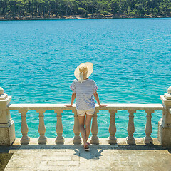 Image showing Rear view of woman wearing straw summer hat ,leaning against elegant old stone fence of coastal villa, relaxing while looking at blue Adriatic sea, on Losinj island Croatia.