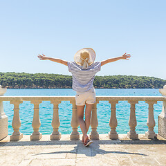 Image showing Rear view of happy woman on vacation, wearing straw summer hat ,standing on luxury elegant old stone balcony of coastal villa, relaxing, arms rised to the sun, looking at blue Adriatic sea