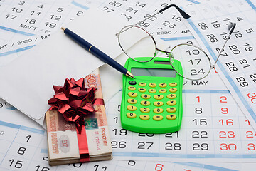 Image showing They put a wad of money in an envelope, rewound with a red ribbon and a bow, next to it lies a calculator, a pen, glasses and a background from calendars