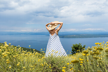 Image showing Rear view of young woman wearing striped summer dress and straw hat standing in super bloom of wildflowers, relaxing while enjoing beautiful view of Adriatic sea nature, Croatia