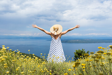 Image showing Rear view of young woman wearing striped summer dress and straw hat standing in super bloom of wildflowers, relaxing with hands up to the sky, enjoing beautiful view of Adriatic sea nature, Croatia