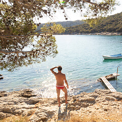 Image showing Rear view of man wearing red speedos tanning and realaxing on wild cove of Adriatic sea on a beach in shade of pine tree.