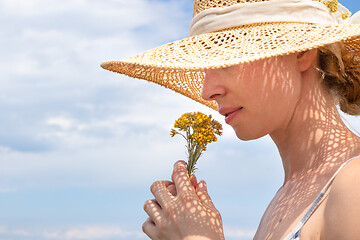 Image showing Portrait of young beautiful cheerful woman wearing straw sun hat, smelling small bouquet of yellow wild florets, against blue summer sky