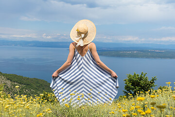 Image showing Rear view of young woman wearing striped summer dress and straw hat standing in super bloom of wildflowers, relaxing while enjoing beautiful view of Adriatic sea nature, Croatia